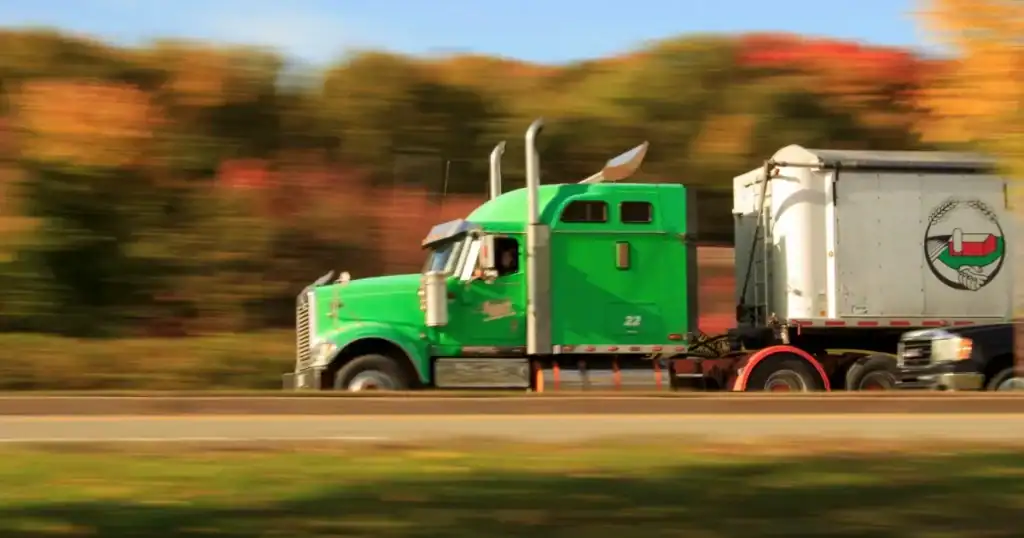 Green semi-truck driving on highway with autumn trees in background.